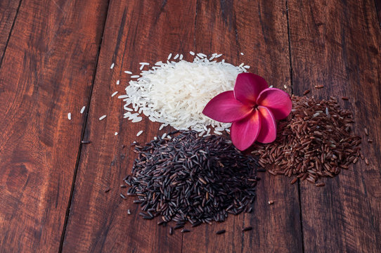 Few heap of different rice on wooden desk. Jasmine white, black and brown rice on kitchen table with Pink plumeria flower