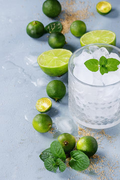 Ingredients for mojito cocktail, whole, sliced lime and mini limes, mint leaves, brown crystal sugar over gray stone texture background with glass full of ice. Space for text
