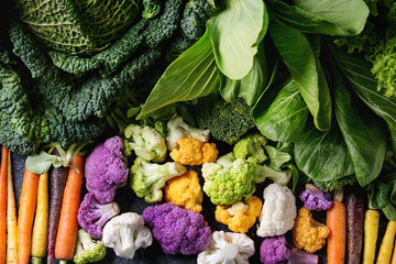 Variety of raw green vegetables salads, lettuce, bok choy, corn, broccoli, savoy cabbage, colorful young carrots and cauliflower over black stone texture background. Top view, space for text