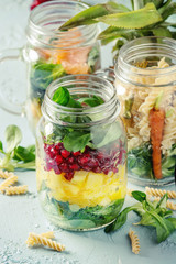 Variety of salads in mason jars. Fruit salad mango, pomegranate, greens, vegetables, wholegrain pasta, carrots and cauliflower, salmon. Standing over blue texture background. Food to go.