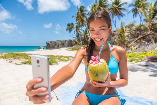 Happy girl taking selfie on summer beach vacation. Cute Asian multiracial bikini woman drinking fresh coconut water smiling holding mobile phone for self-portrait picture on tropical travel.