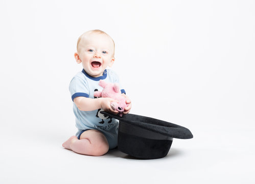 Baby Holding Stuffed Animal Pulled Out of Black Hat. a 6 month old baby kneeling on white finds a stuffed animal pig is inside a black fedora hat and smiles at camera