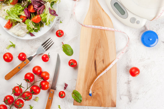 Concept of weight loss and healthy eating: a light salad of green leaves and tomatoes, centimeter tape and scales. On a white concrete table, copy space top view