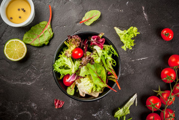 Light salad of green leaves and tomatoes, oil and lime juice. On a black concrete table, with ingredients, copy space top view