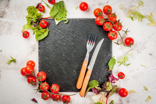 Cooking, healthy food: fresh salad leaves and tomatoes on the table, with a black cutting board, cutlery. Top view, on white concrete background