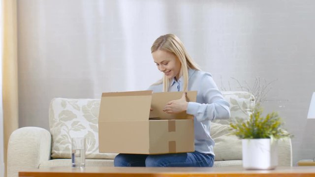Beautiful Young Woman at Home Opens Cardboard Box while Sitting on a Couch in Her Living Room.  Shot on RED EPIC-W 8K Helium Cinema Camera.