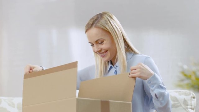Beautiful Young Woman at Home Opens Cardboard Box while Sitting on a Couch in Her Bright Living Room. She Smiles.  Shot on RED EPIC-W 8K Helium Cinema Camera.