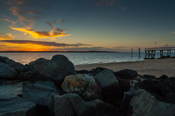 Naklejka premium Large stones in the foreground with a rocky beach, a wooden dock, and a colorful sunset sky with clouds in the background. West Neck Beach in Huntington, NY, USA.