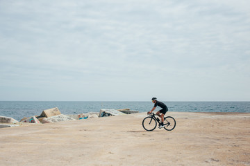 Fototapeta premium Male professional cyclist rides around a pier wearing all black near blue sea and rocky port nice light.