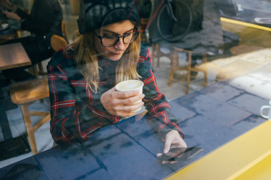 Young Woman Drinks Coffee And Plays On Phone Wearing Flannel Shirt And Beanie Hat Photo Through Window