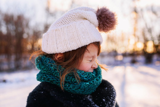 Close-up of a young girl wrapped up in a winter coat, woolly bobble hat and scarf standing in a snowy garden pulling funny faces