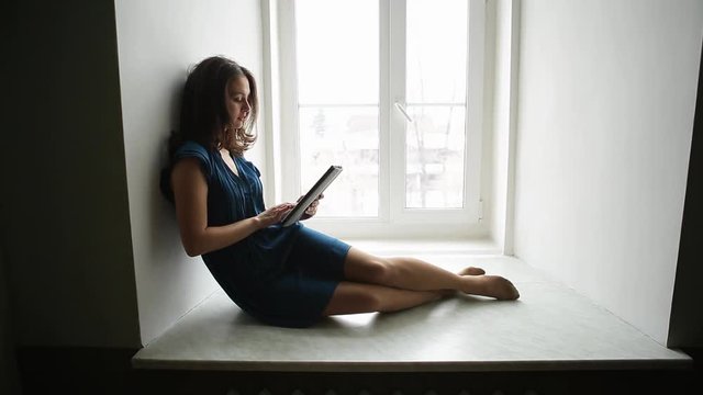 Prores. Young girl in a blue dress is sitting with a tablet on the window