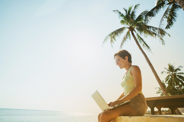 Woman freelancer working behind laptop sitting on the beach under a palm tree. Dream work.
