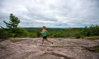 A young woman wearing fitness clothing running across rocks with forest background