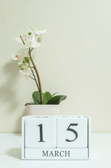 Closeup white wooden calendar with black 15 march word with white orchid flower on white wood desk and cream color wallpaper in room textured background , selective focus at the calendar