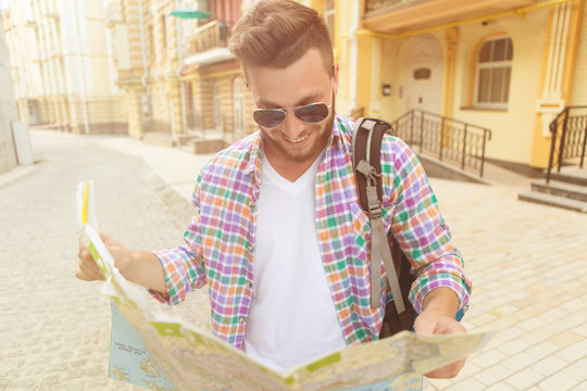 Attractive hipster man in sunglasses looking at city map. Man in plaid shirt with backpack looking for directions. Toned image.
