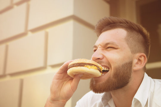 Man eating hot hamburger after stressful work. Close-up of hipster bearded man eating delicious hamburger outdoors. Toned image.