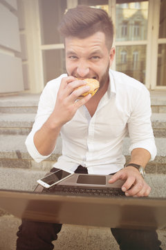 Handsome man freelancer working on laptop and eating hamburger. Bearded man in white shirt and black jeans looking at the camera. Toned image.