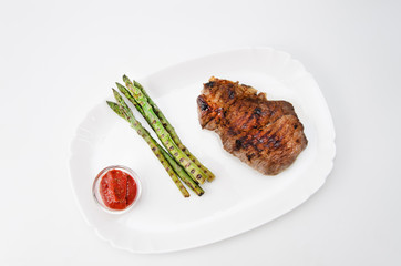 Grilled beef steak with asparagus and red sauce on a white plate, over white background