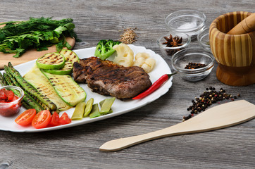 Grilled beef grilled with asparagus, zucchini, lobi, broccoli, tomato, chili and red sauce on a white plate, and other vegetables in wood background