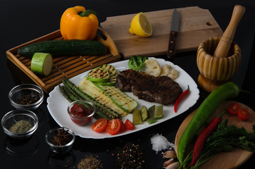 Grilled beef grilled steak with asparagus, zucchini, lobi, broccoli, tomato, chili and red sauce on a white plate, on a black glossy background