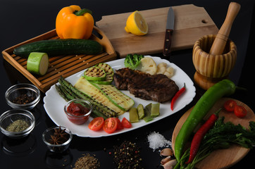 Grilled beef grilled steak with asparagus, zucchini, lobi, broccoli, tomato, chili and red sauce on a white plate, on a black glossy background