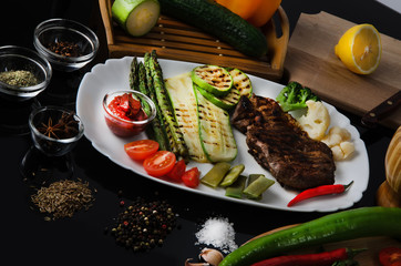 Grilled beef grilled steak with asparagus, zucchini, lobi, broccoli, tomato, chili and red sauce on a white plate, on a black glossy background
