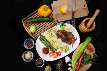 Grilled beef grilled steak with asparagus, zucchini, lobi, broccoli, tomato, chili and red sauce on a white plate, on a black glossy background