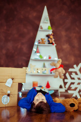 Boy in blue Christmas sweater lies with present boxes on wooden floor in cosy room