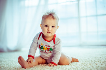 Little child in white baby-sleeper with lettering 'Santa' sits on fluffy carpet