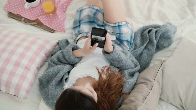 Young beautiful woman lying on the bed and holding mobile phone. Girl watches photos on smartphone and drinks juice.