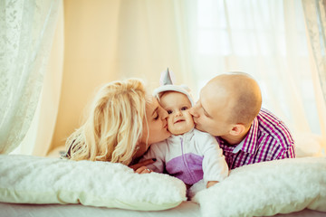 Fototapeta na wymiar Joyful parents kiss their little daughter lying on soft bed