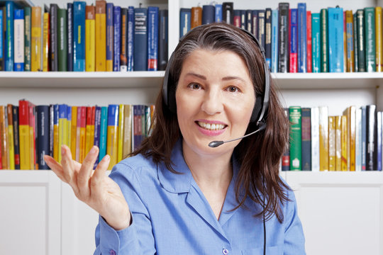 Smiling woman with headset in a library, explaining something during a video call, telelearning concept.