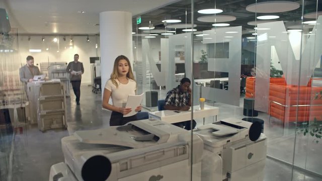 Lockdown of people working in modern office with glass walls: attractive Asian businesswoman putting paper into printer, African businessman and Caucasian male colleagues working in background 