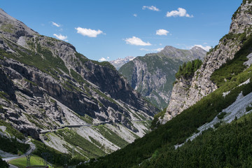 Aerial view of alpine road in valley between two high mountains coated with green forests. Blue sky with clouds on background.
