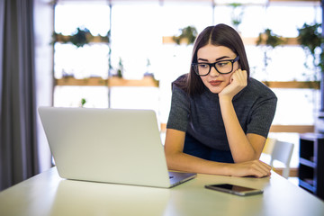 Cheerful young beautiful woman in glasses looking at laptop with smile while sitting at her working place