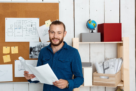 Young successful architector smiling, holding drawings, standing in office background.