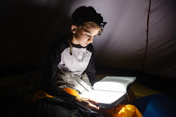 Teen boy sitting in a camping tent wrapped in a sleeping bag reading a book with flashlight on at...