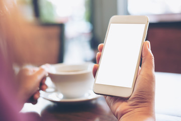 Mockup image of hands holding white mobile phone with blank white screen and hot coffee cup in vintage loft cafe