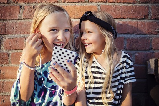 Happy girls listening music through smart phone while sitting against brick wall