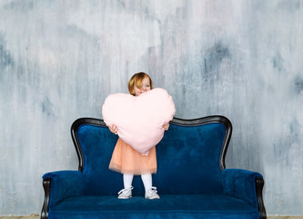 The cute baby girl stand on a sofa and holds heart pillow in hands.