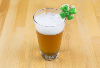 Amber beer drink in a transparent glass with a high white foam and clover on a wooden base. The day of St. Patrick's Day.