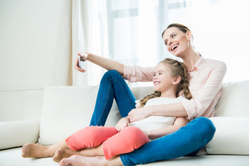 side view of happy mother and daughter sitting on sofa and making selfie