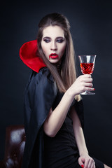 The girl is a vampire in a cloak dracula with a glass in her hand.