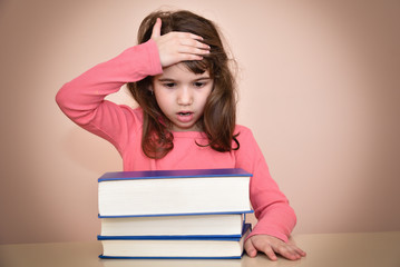 Surprised and astonished young girl holding her head and sitting at the table with books wondering how much she has to read