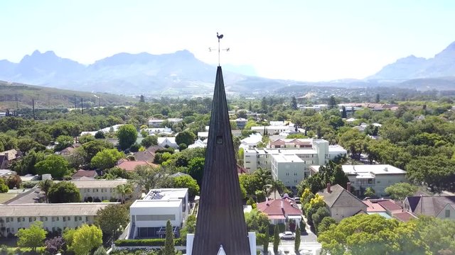 Aerial of white church moving showing small city town and mountains in background then moving more backwards and down revealing a white church with darker tower and cross on top of tower 4k quality