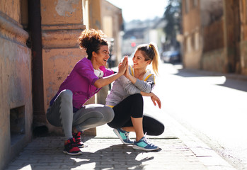 Two young woman taking a break after jogging ,early in the morning.They crouching and laughing.