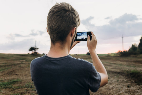 stylish hipster traveler holding smart phone taking photo of beautiful sunset landscape in summer field. instagram photography. exploring and discovering. space for text.