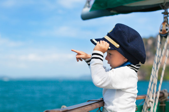 Funny little baby captain on board of sailing yacht watching offshore sea on summer cruise. Travel adventure, yachting with child on family vacation. Kid clothing in sailor style, nautical fashion.