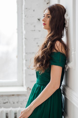 beautiful brunette girl with long hair standing in green dress near the white wall by the window looking to the side